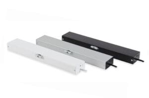 Three rectangular metal cable management boxes are arranged in a row, featuring hinged lids and side openings for cable entry. The boxes, ideal for integrating window actuators or door automation systems, are colored white, silver, and black respectively.