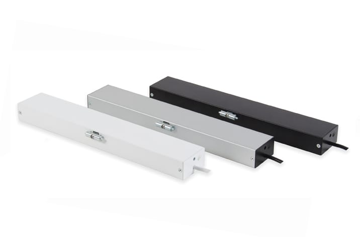 Three rectangular metal cable management boxes are arranged in a row, featuring hinged lids and side openings for cable entry. The boxes, ideal for integrating window actuators or door automation systems, are colored white, silver, and black respectively.
