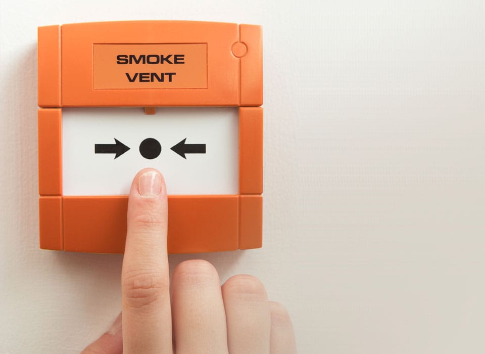 A close-up of a finger pressing a button on an orange smoke vent activation panel mounted on a white wall, seamlessly integrated with the building's door automation system.