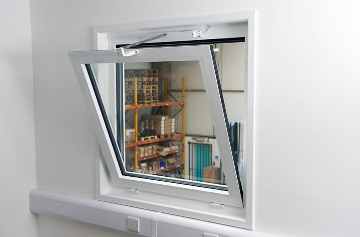Mingardi Micro Evo1 actuator fitted to an inward opening bottom hung window. small white-framed awning window is slightly open, revealing a view of a storage room with shelves and stacked items, possibly equipped with window actuators for automatic opening and closing.