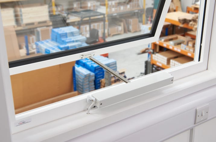Mingardi Micro Evo1 Chain Actuator fitted to a window. A mechanical chain window opener and control unit, equipped with advanced window actuators, is installed on a white window, overlooking a room filled with blue boxes and various stored items. This setup not only ensures natural ventilation but also contributes to an efficient smoke ventilation system.
