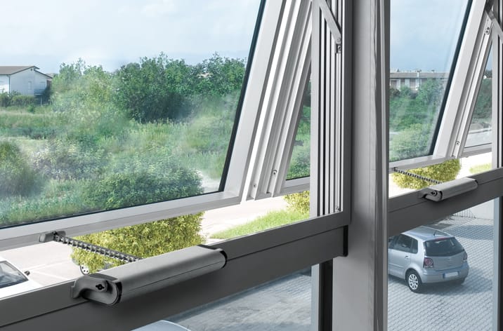 A row of windows in a building are open outward, revealing a view of parked cars and greenery outside. The windows are equipped with mechanical arm mechanisms, showcasing advanced window actuators for seamless operation.