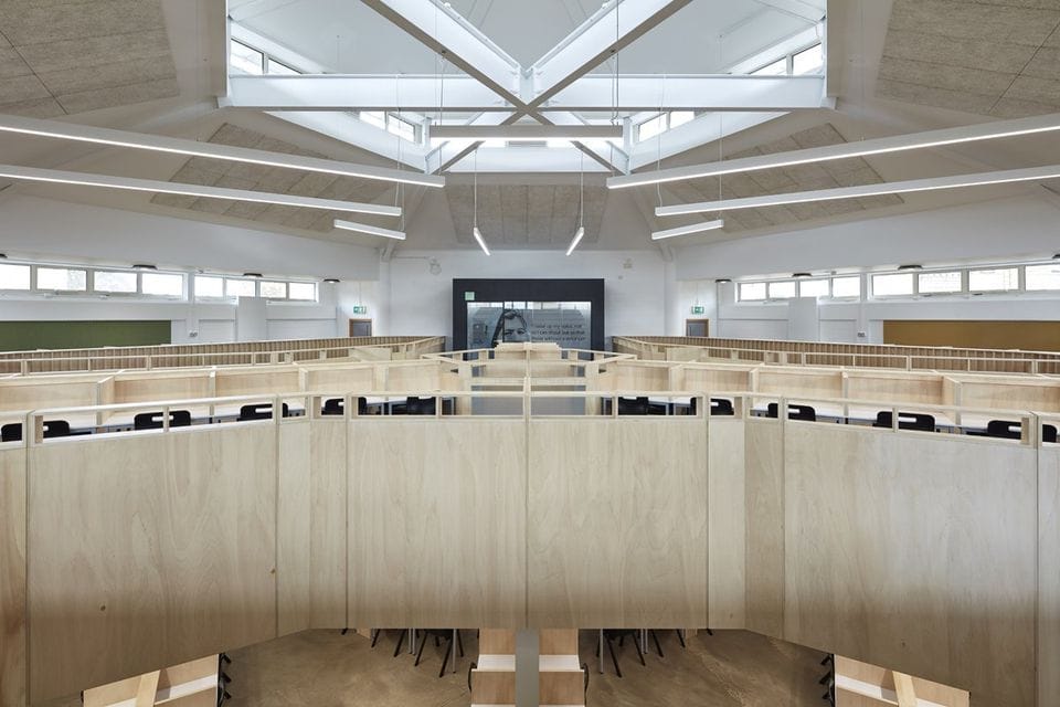 Wide-angle view of an empty, modern lecture hall with tiered wooden seating, a large skylight, and a podium in the center. Numerous windows line the upper part of the walls.