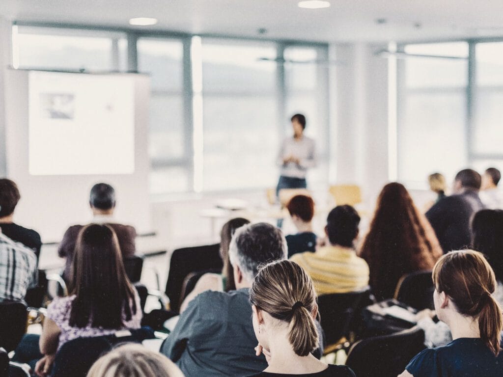 A group of people seated in a classroom setting, facing a presenter standing near a screen displaying a presentation.