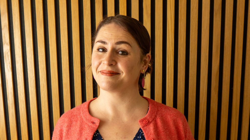 A person with brown hair and a pink cardigan smiles slightly, standing in front of a wall with vertical wooden slats.