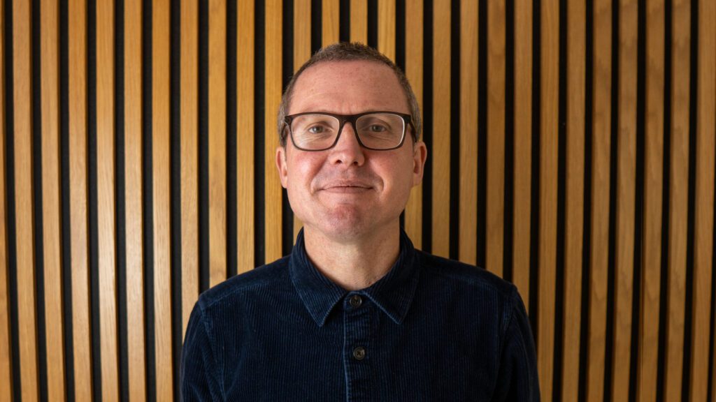 A person wearing glasses and a dark shirt stands in front of a wall with vertical wooden slats.