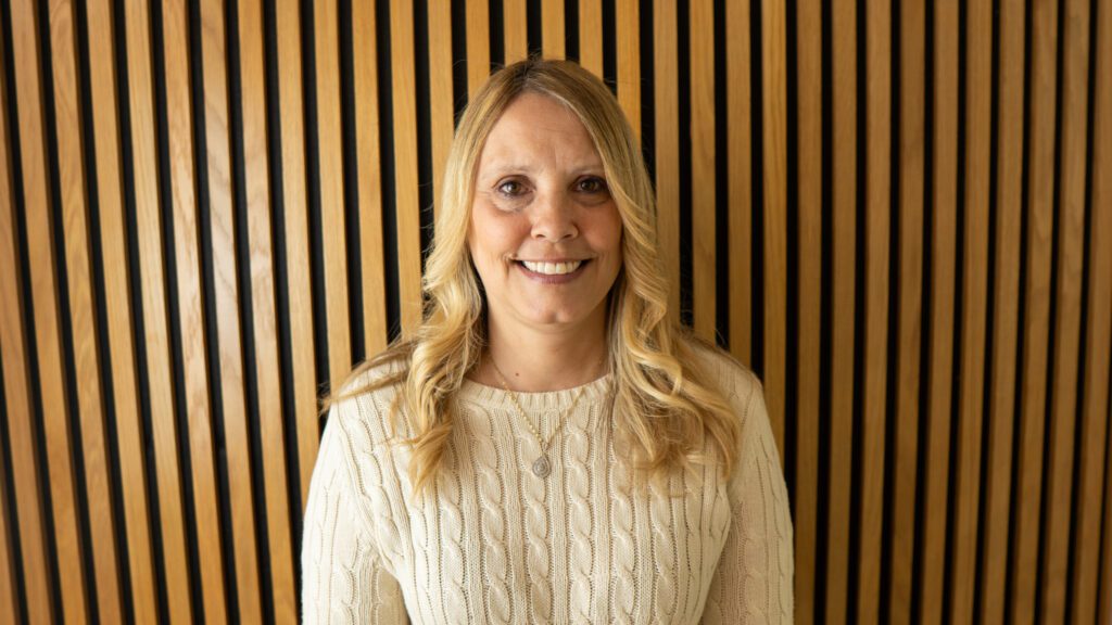 A woman with long blonde hair wearing a cream cable-knit jumper stands in front of a wall with vertical wooden slats.