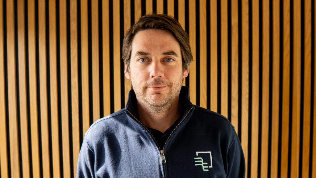 A man with brown hair wearing a navy zip-up jacket stands in front of a wall with vertical wooden battens.