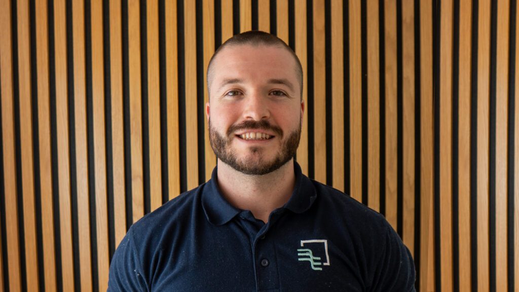 A man with a beard in a navy polo shirt stands in front of a vertical wooden slat wall, smiling at the camera.