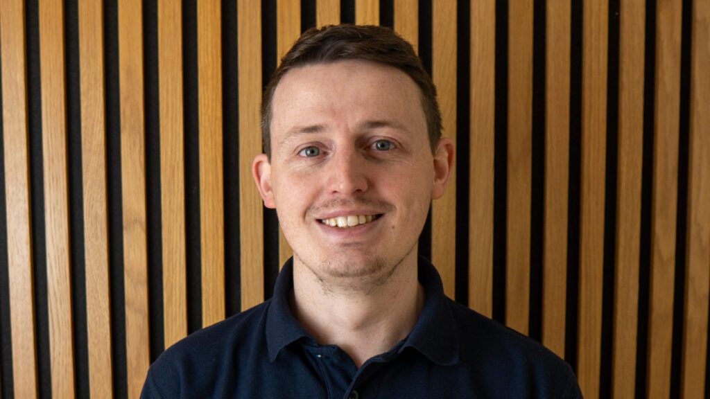 A man with short brown hair and a navy collared shirt stands in front of a wooden slat wall, smiling at the camera.