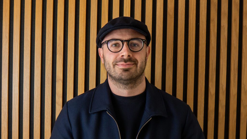 A man wearing glasses, a black cap, and a dark jacket stands in front of a wall with vertical wooden slats.