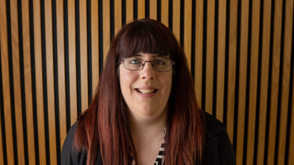 A person with long dark hair and glasses is smiling in front of a wall with vertical wooden battens.
