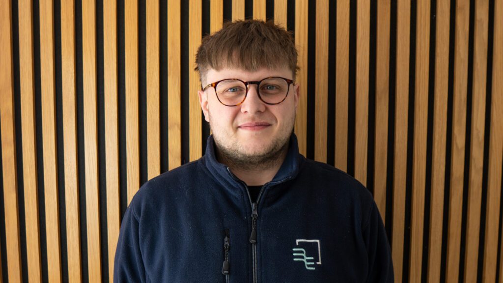 A person with short hair, glasses, and a navy jacket stands in front of a wall with vertical wooden slats.