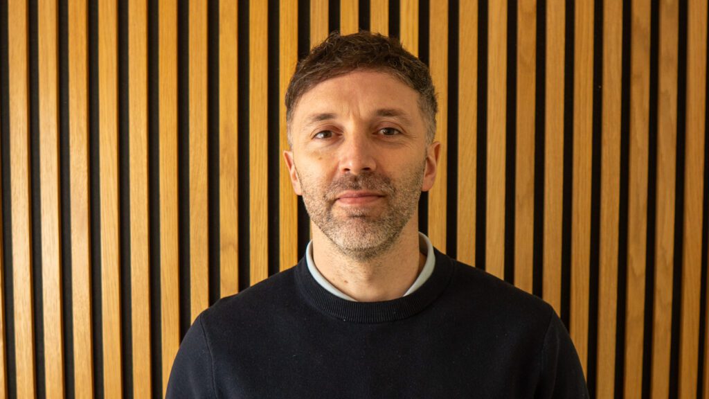 A man with short brown hair and a trimmed beard stands in front of a wall with vertical wooden slats, wearing a dark jumper over a collared shirt.