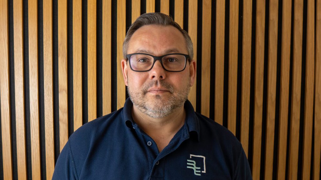 A man with glasses and short hair stands in front of a wall with vertical wooden slats, wearing a navy blue polo shirt with a white and green logo.