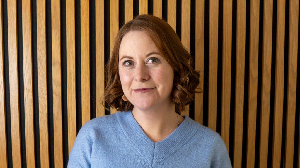 A woman with shoulder-length red hair and a light blue jumper stands in front of a background of vertical wooden slats.